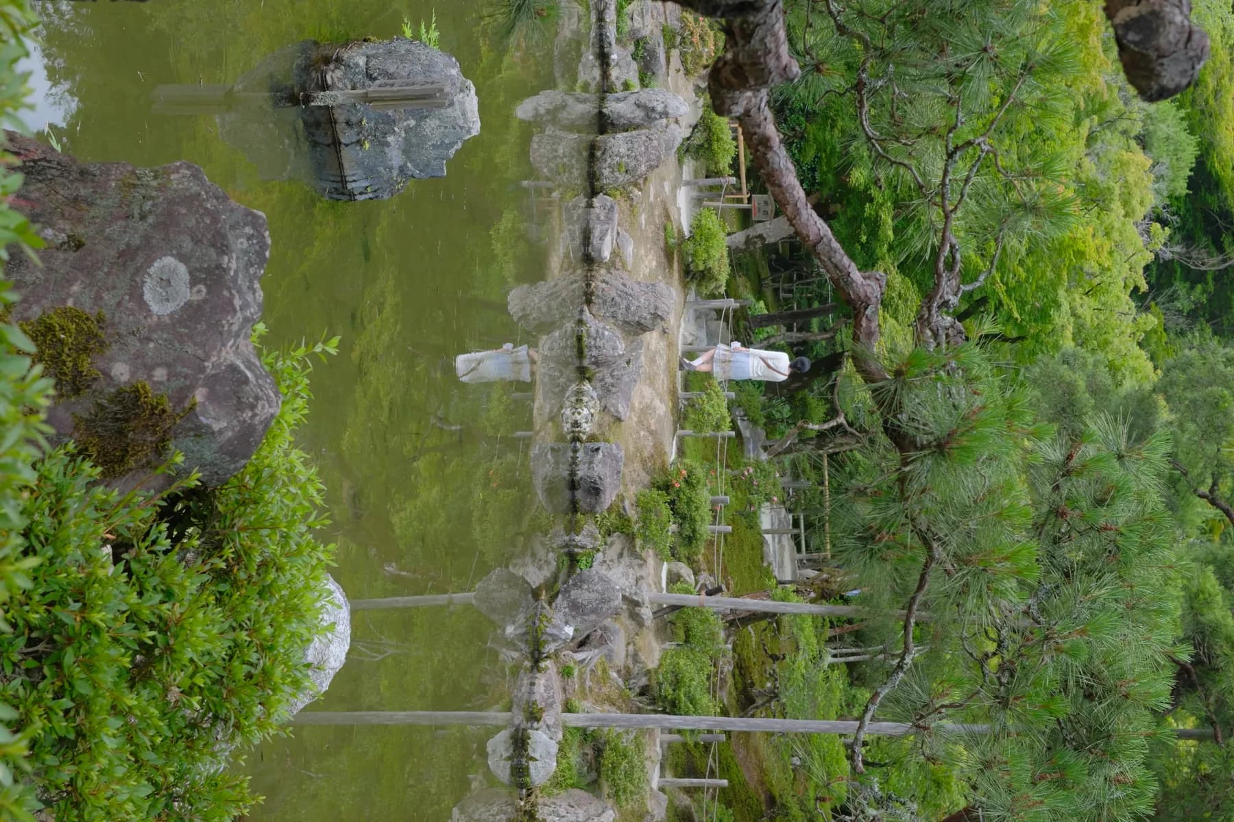 A woman walks through a tranquil Japanese garden with a pond and rocks