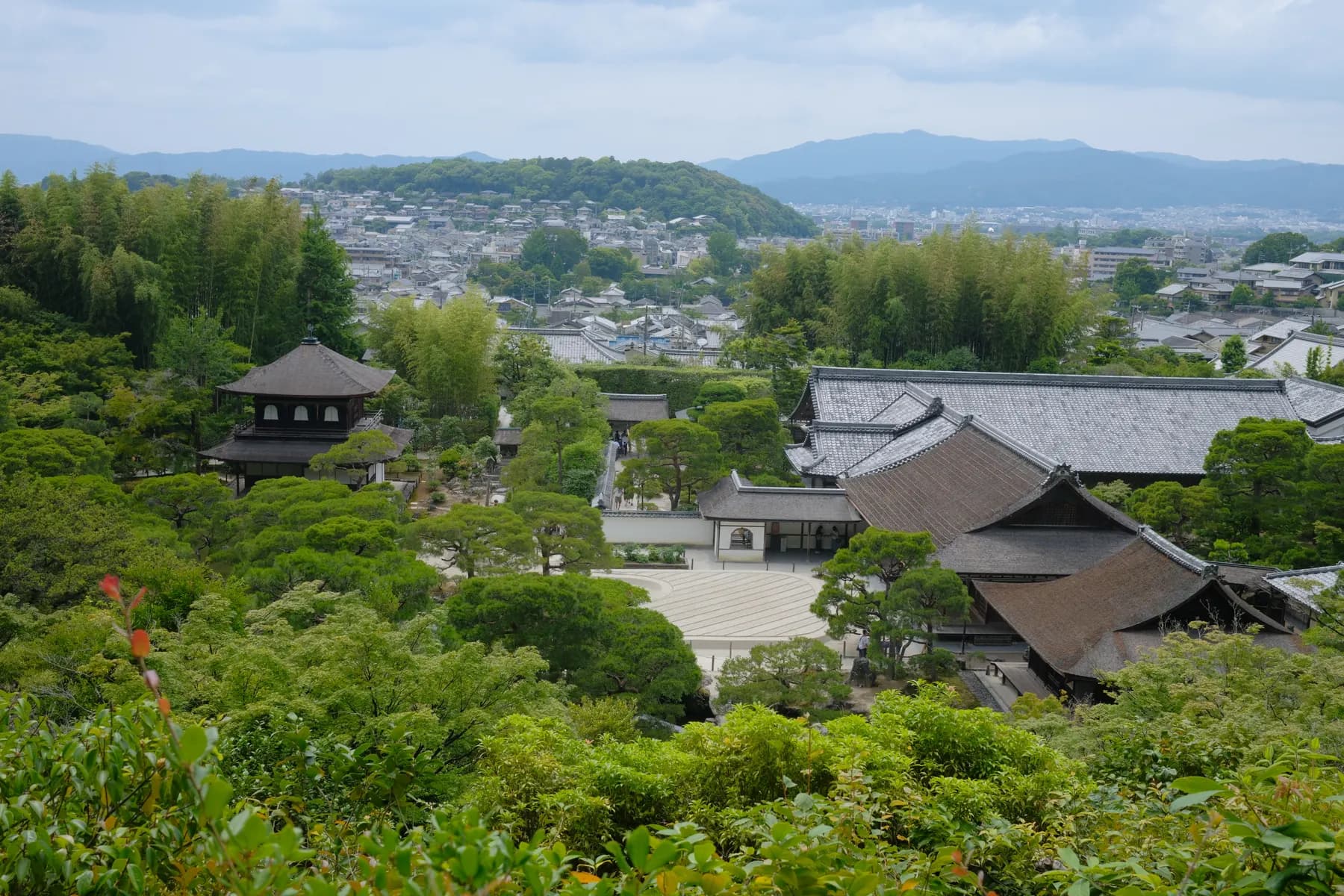A tranquil view of a Japanese temple complex nestled amidst lush greenery, with a distant cityscape and mountains in the background