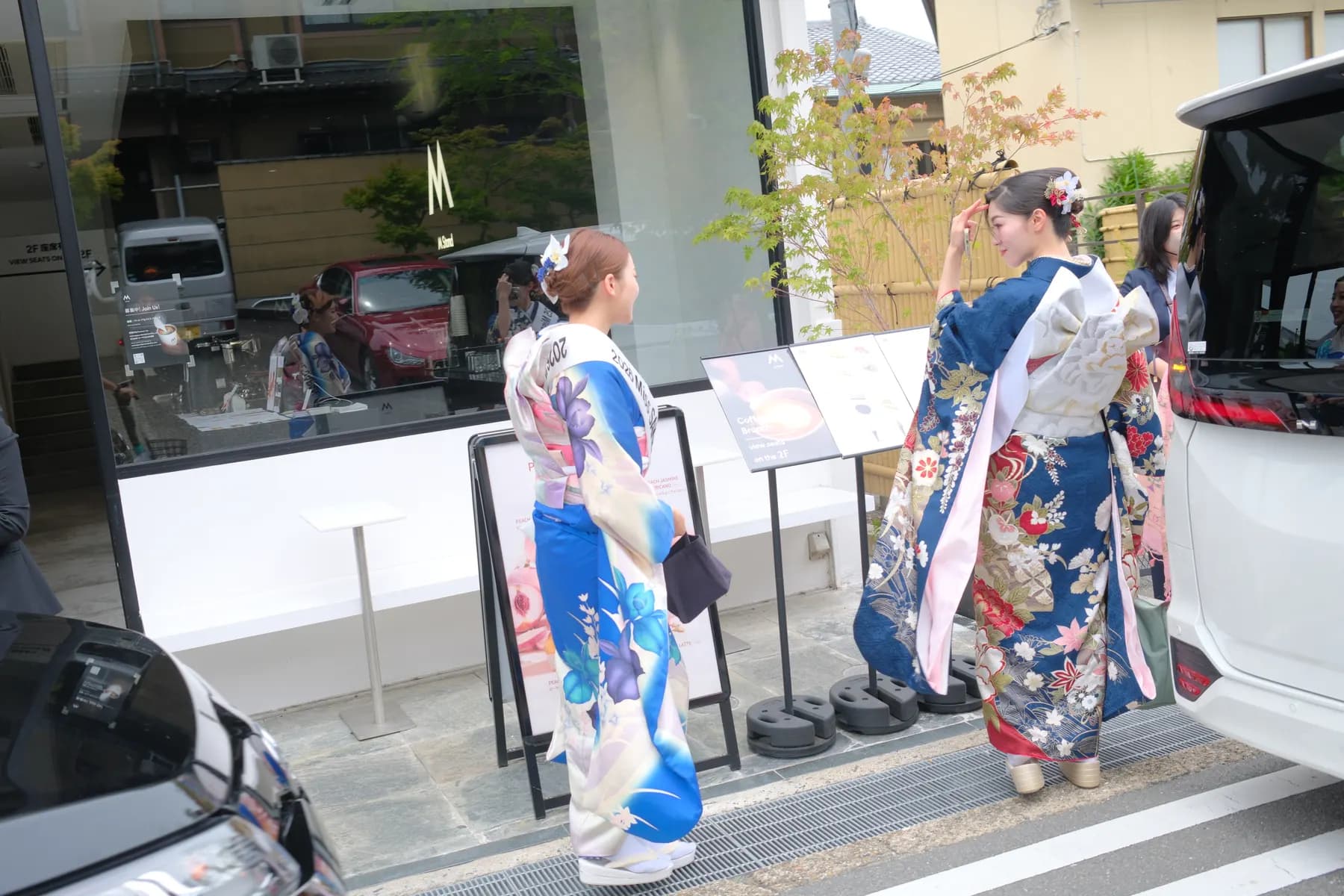 Two women dressed in traditional kimonos are posing for a photo in front of a building with signboards and parked cars