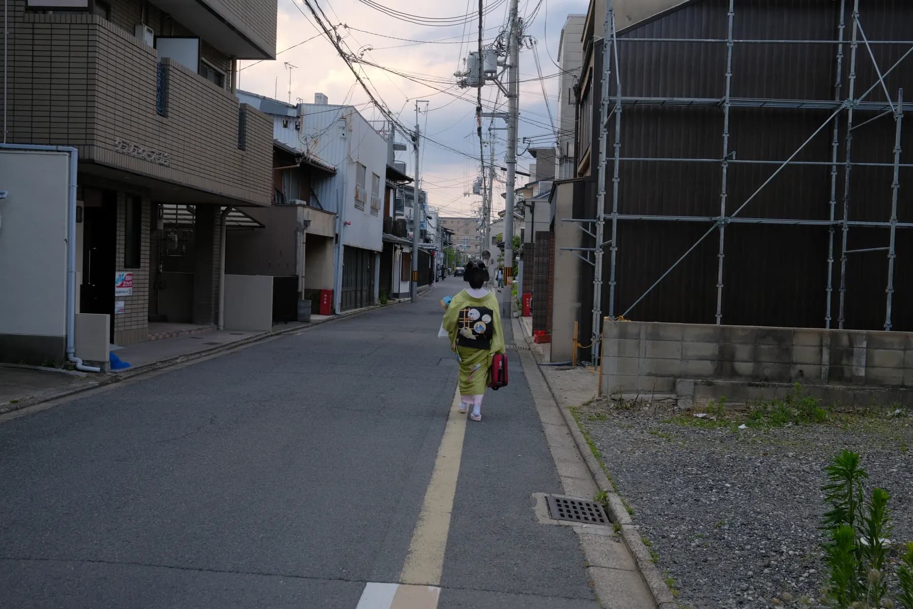 A woman in a traditional kimono walks down a quiet street lined with buildings and scaffolding, under a dusky sky