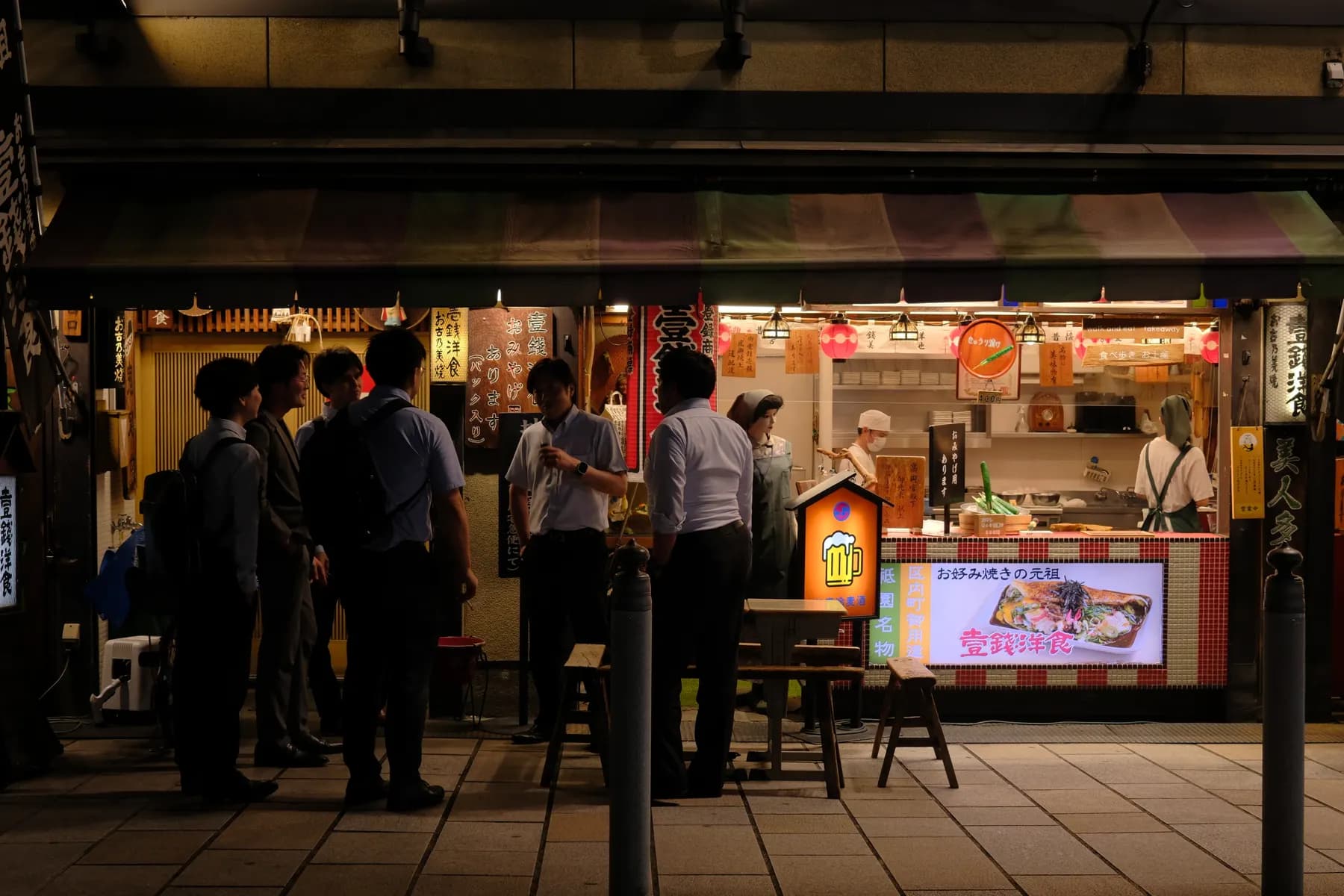A group of people gathered around a food stall at night, illuminated by the warm glow of the stall's lights