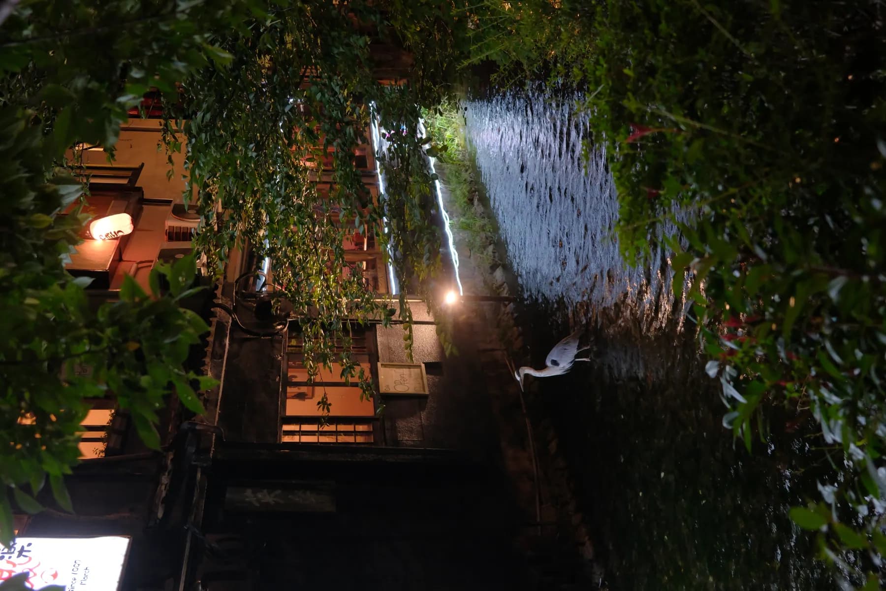 A serene night scene featuring a bird standing in a river surrounded by illuminated buildings and trees