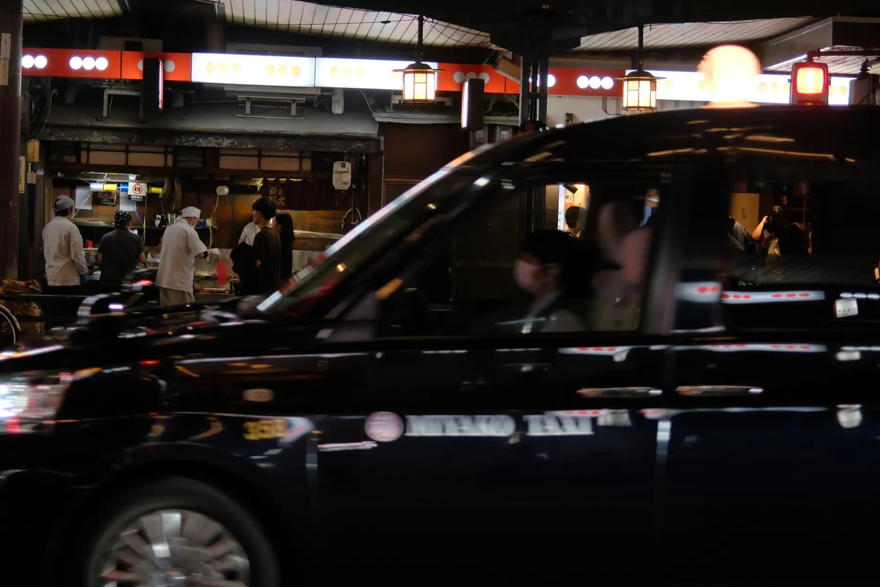 A bustling street scene at night with a taxi in motion and restaurant staff preparing food