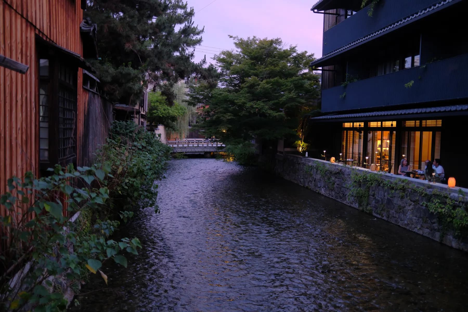 A tranquil scene of a river at sunset, flanked by traditional buildings and lush greenery, with people enjoying the view