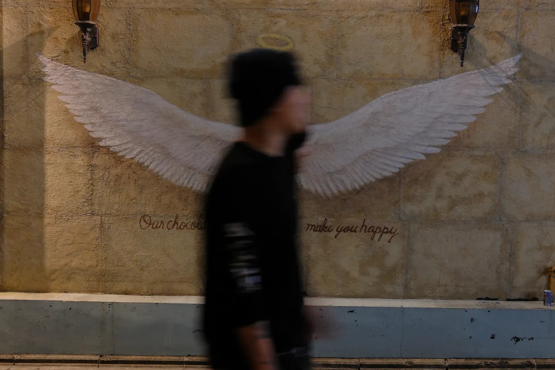 A person walks past a mural of angel wings and a halo on a wall, with the text 'Our chocolate make you happy' below