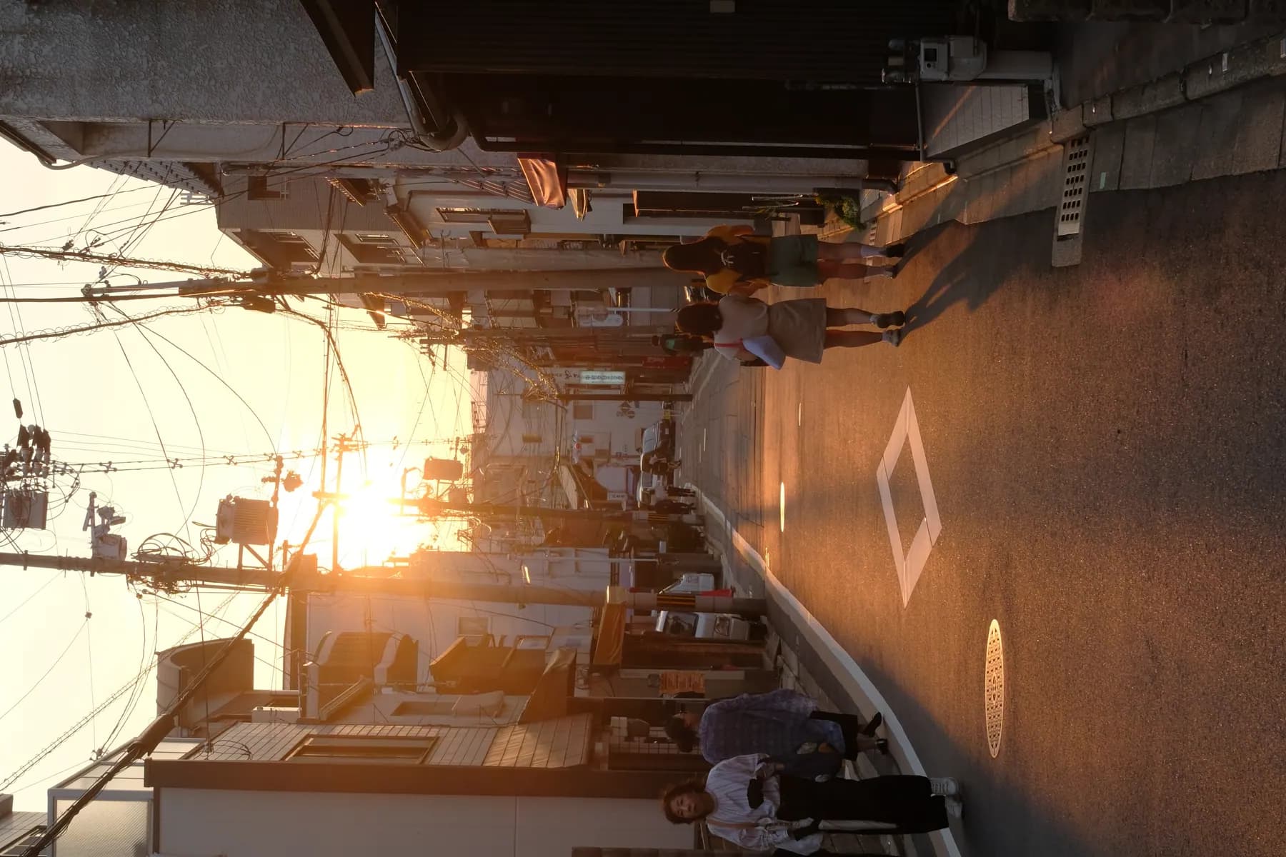 A serene sunset scene in a narrow urban street with people walking and the warm glow of the setting sun casting long shadows