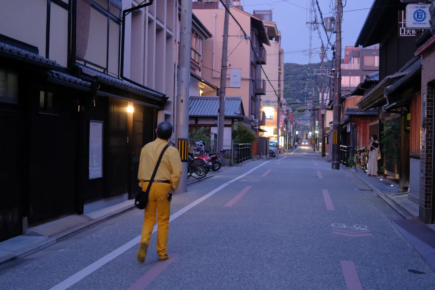 A man in a bright yellow suit walks down a quiet, narrow street lined with traditional buildings in the early evening