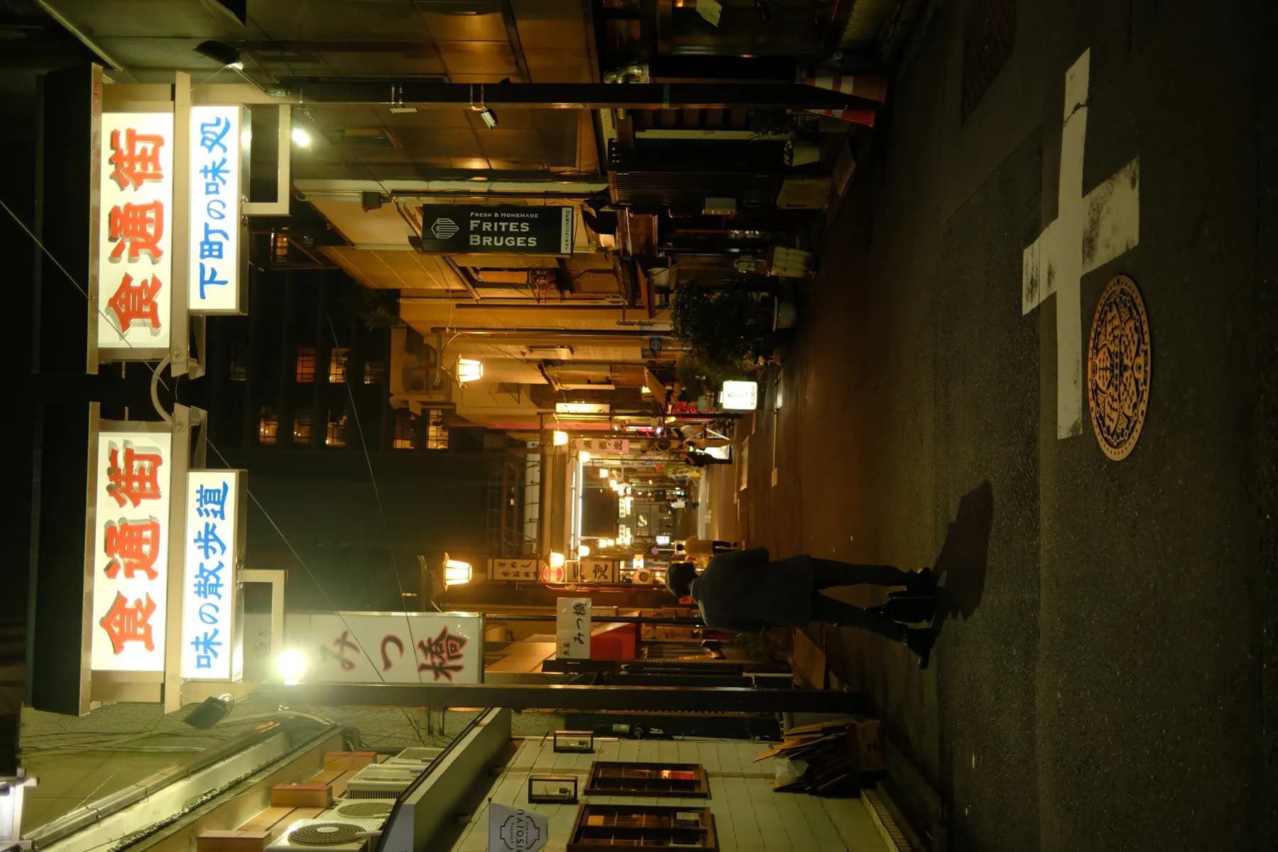 A person walks down a dimly lit alleyway at night, flanked by illuminated signboards and buildings