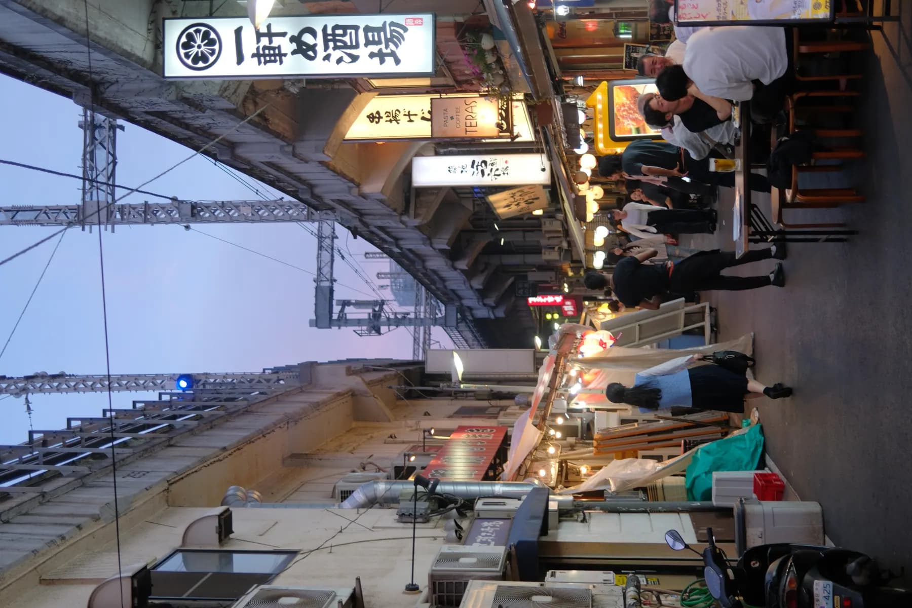 A bustling street scene at sunset with people dining at food stalls and walking along a narrow alleyway lined with shops and restaurants