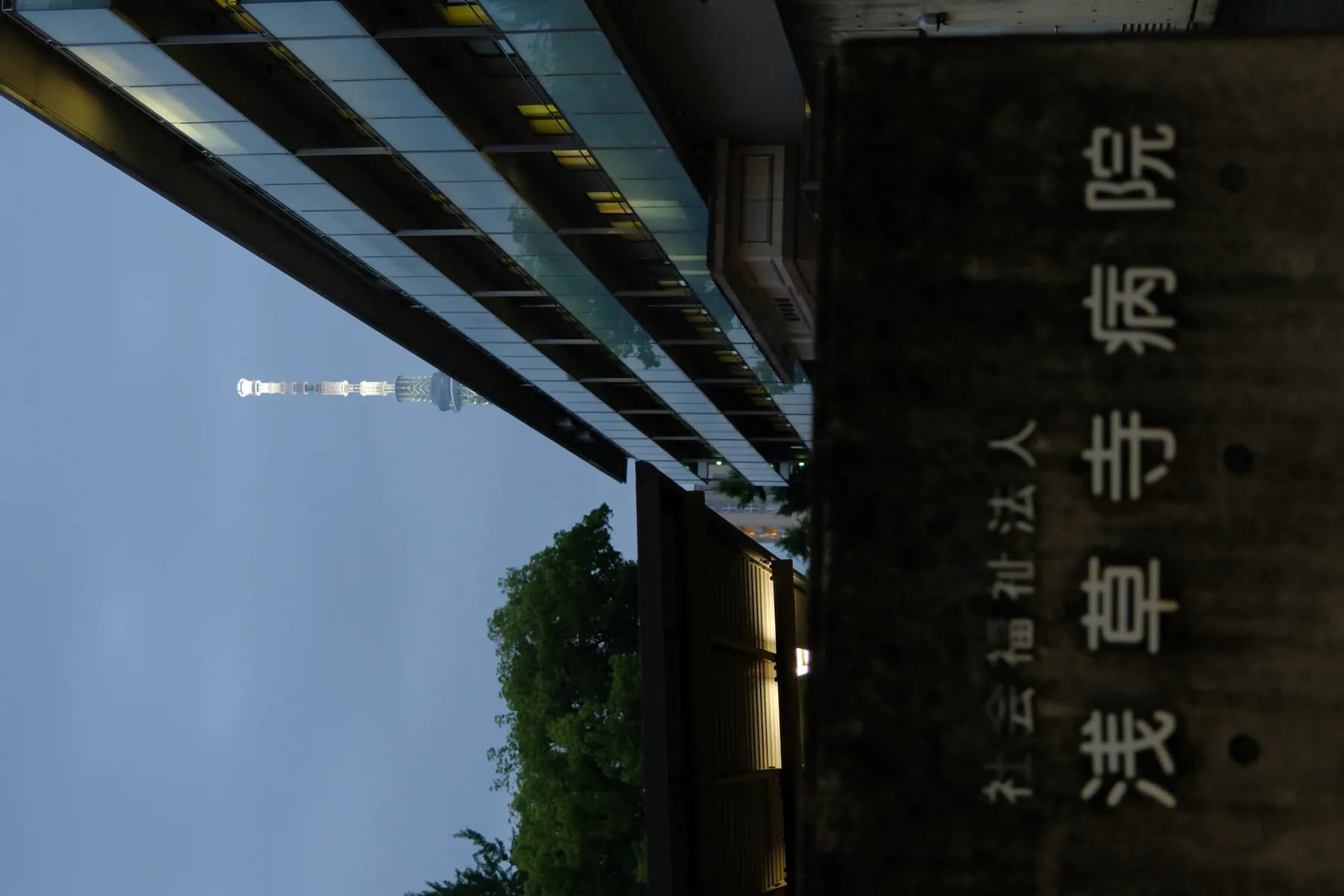 A serene sunset scene featuring a modern building, a tower, trees, and a sign in Japanese, with a calm and peaceful atmosphere