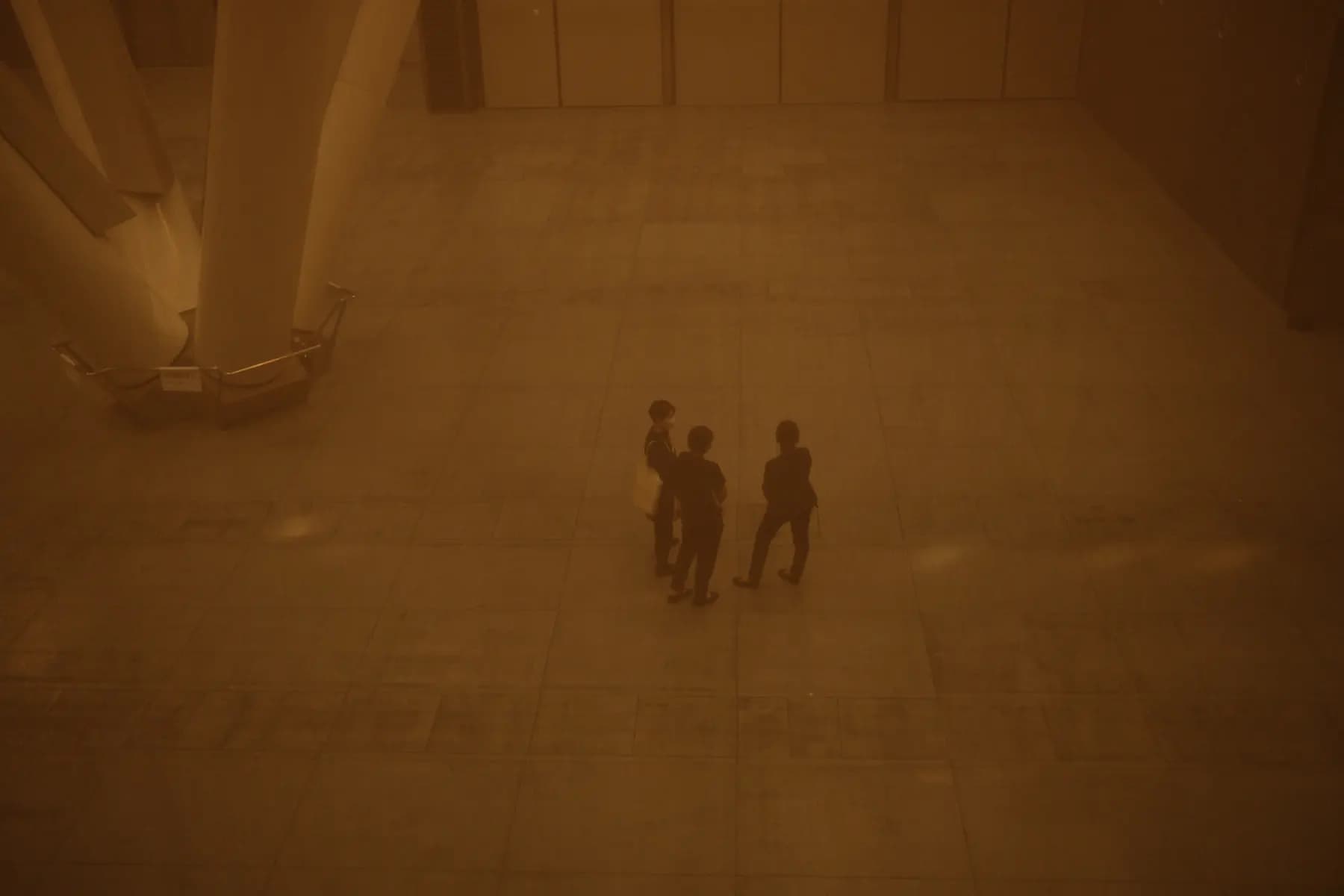 Three people stand in a vast, dimly lit space, surrounded by a large boat and architectural columns, creating a sense of solitude and intrigue