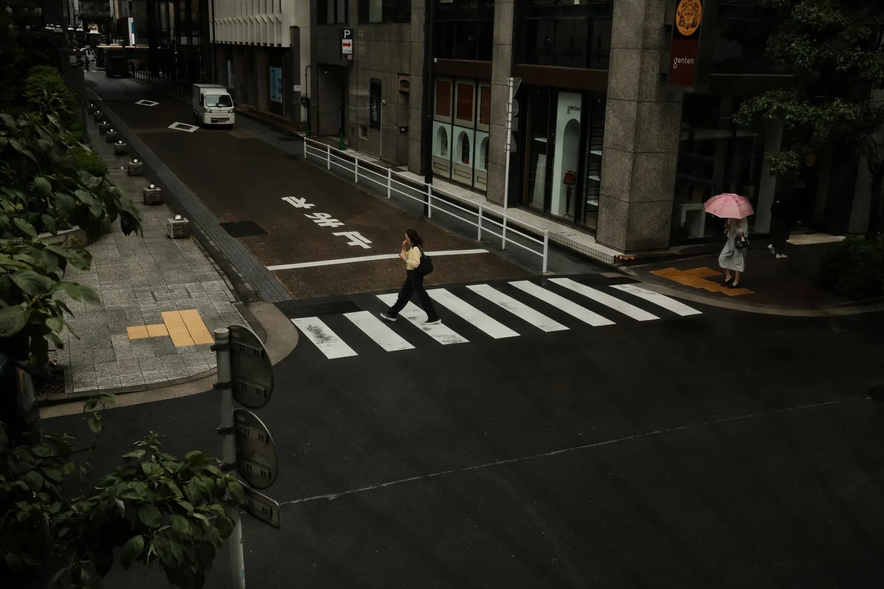 A pedestrian crossing a street in an urban setting with a person walking and another holding an umbrella