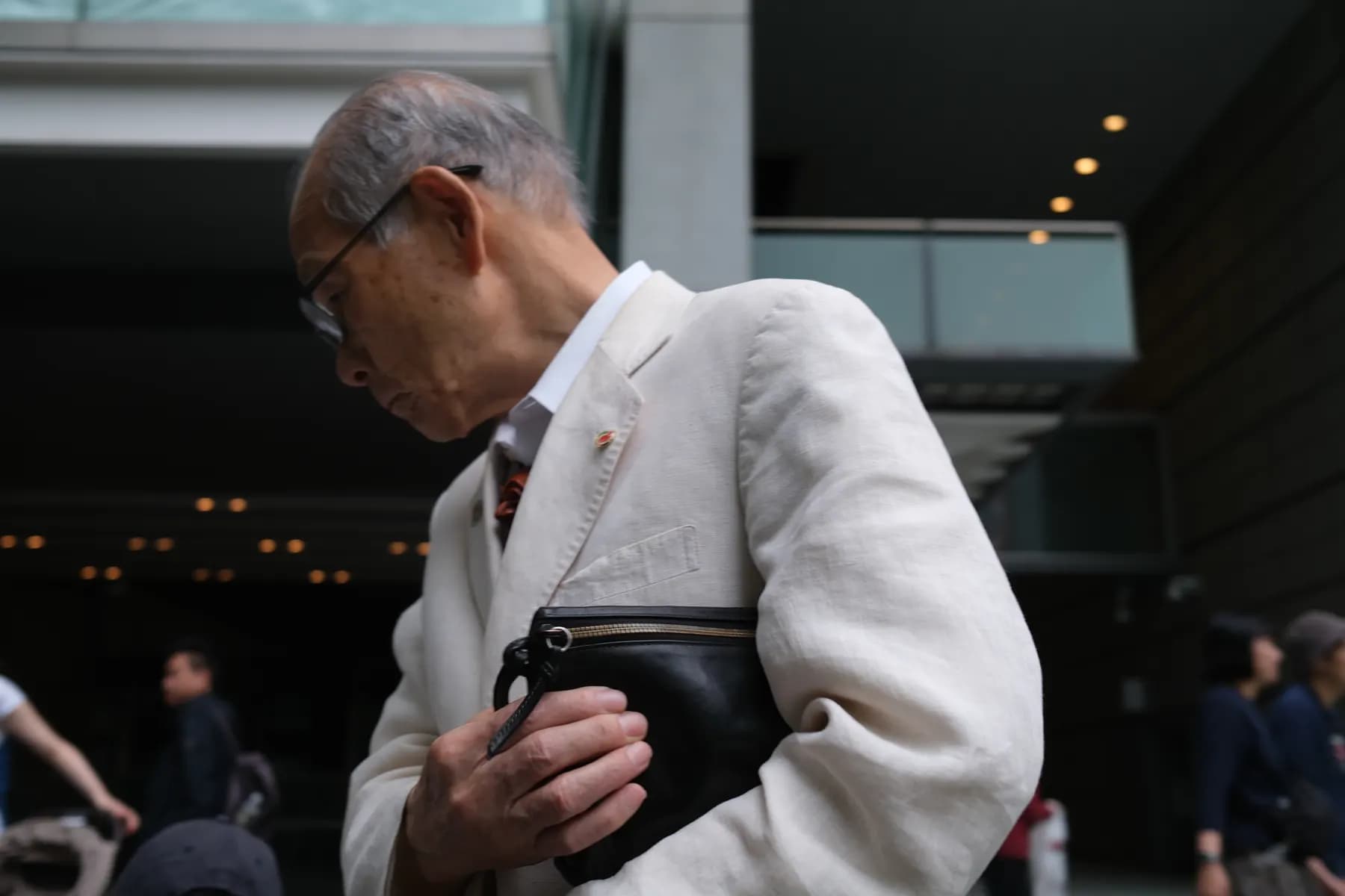An elderly man in a white suit walks through a modern urban setting, holding a black bag
