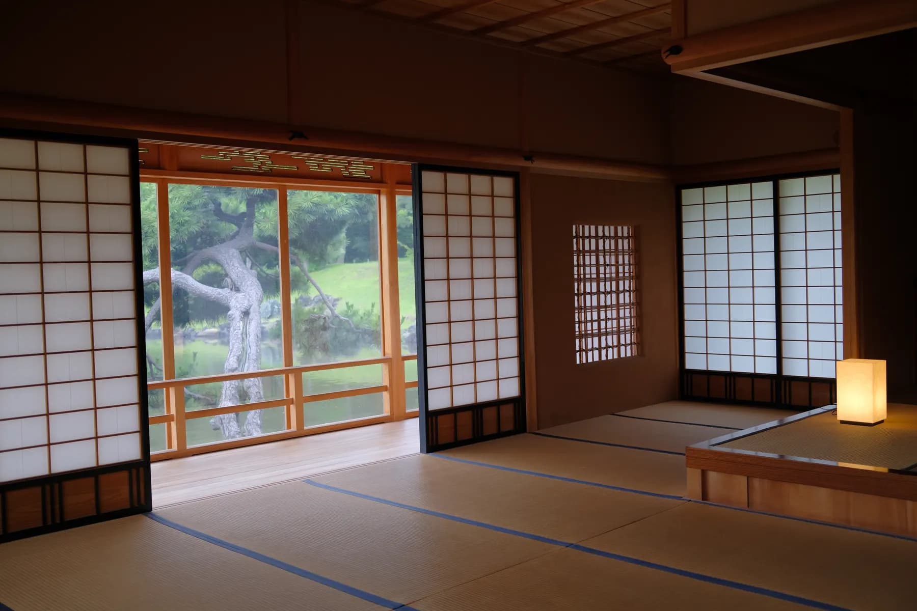 A tranquil Japanese room with sliding shoji screens and tatami mats, bathed in soft light from a floor lamp, overlooking a serene garden