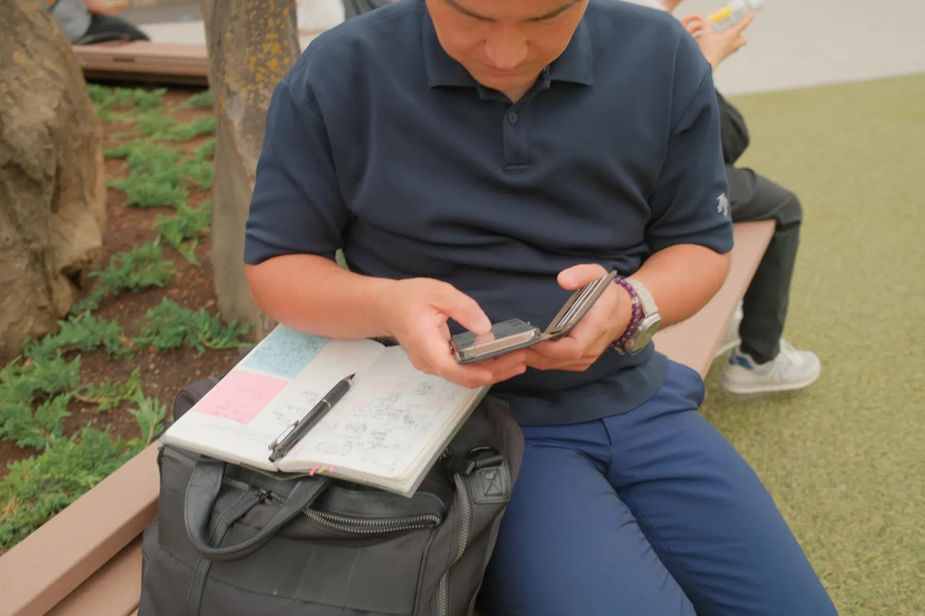 A man is sitting on a bench outdoors, focused on his phone, with a notebook and pencil resting on his bag