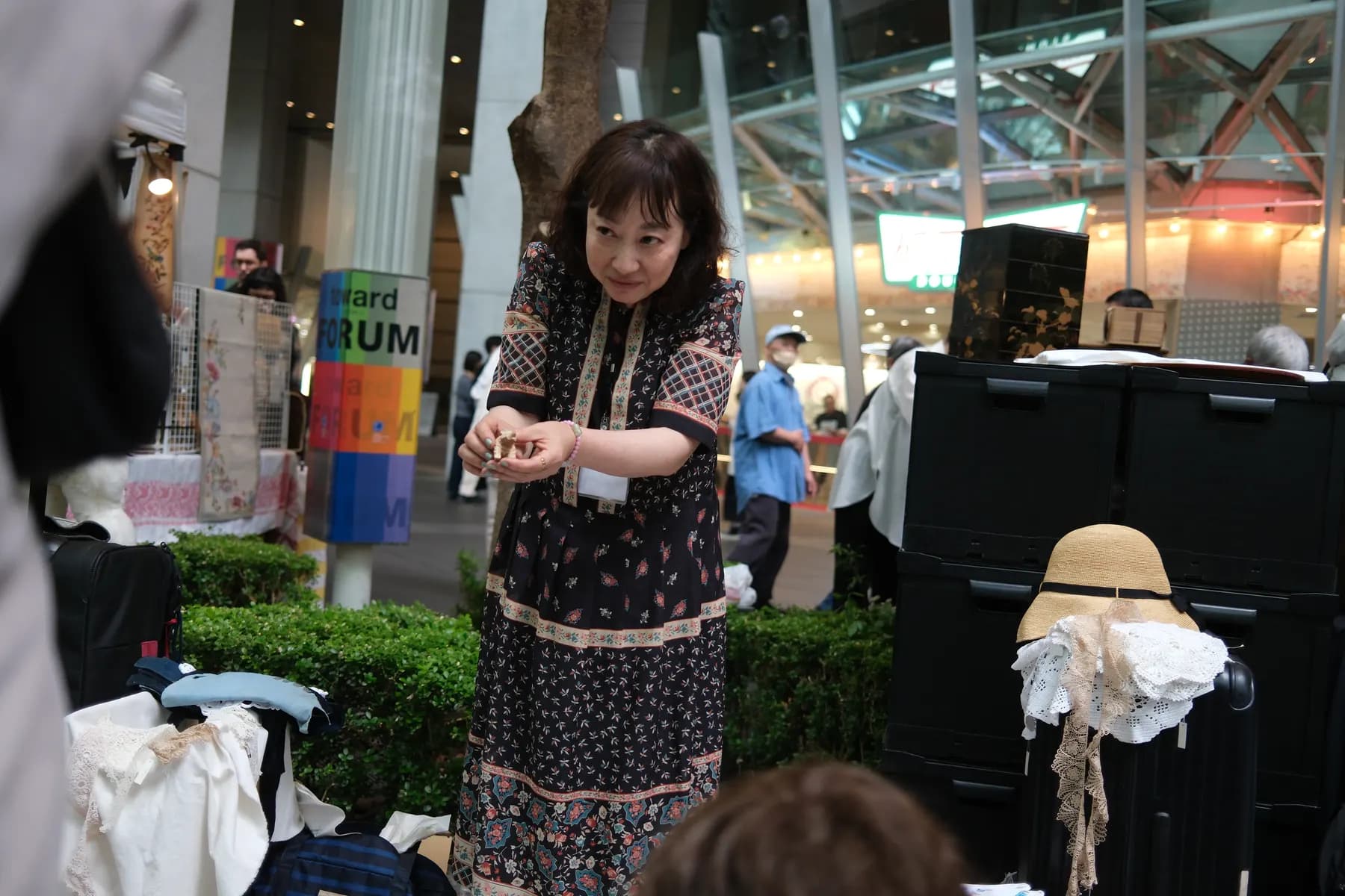 A woman in a floral dress stands amidst a bustling outdoor market scene, surrounded by various objects and people