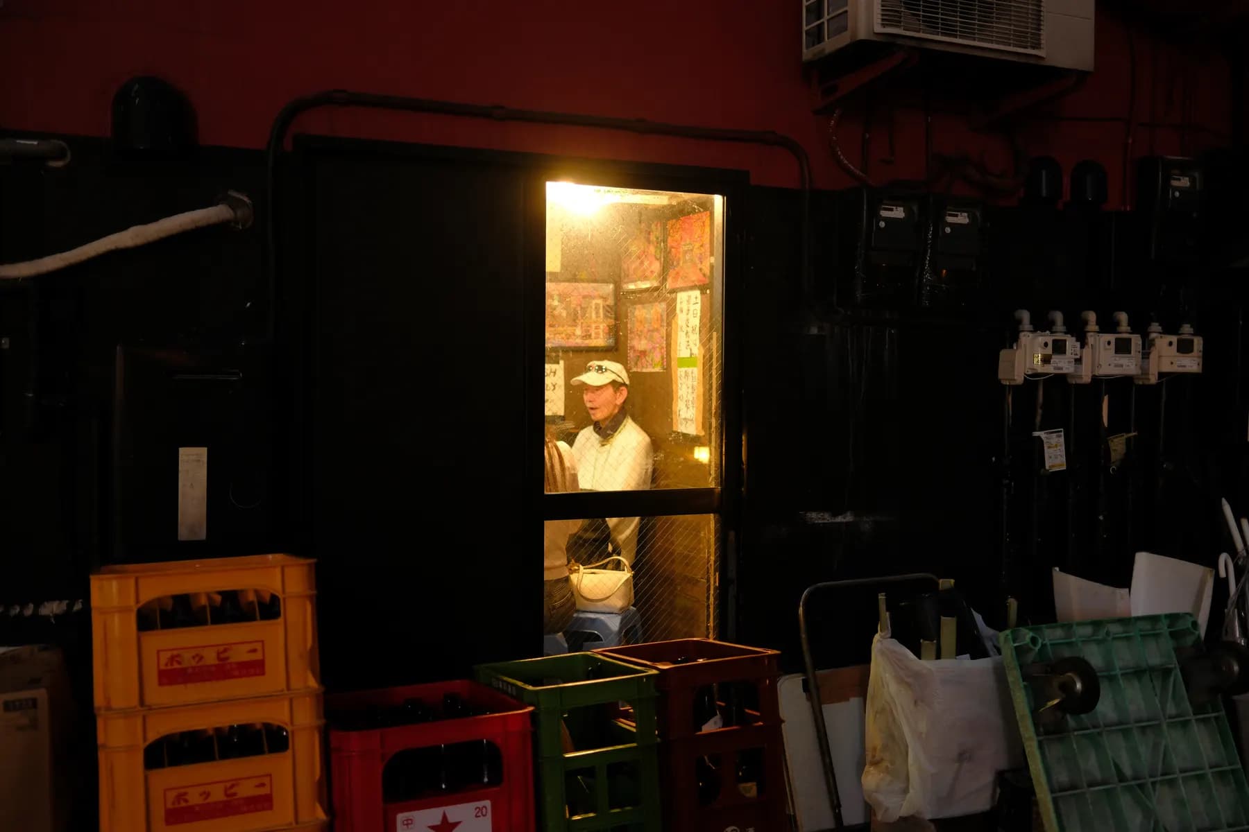A man stands inside a dimly lit room, surrounded by crates and various objects, illuminated by a single light source