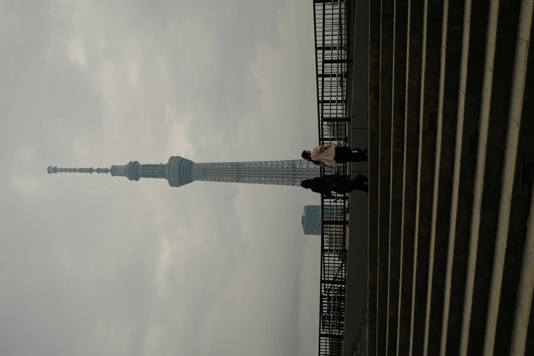 Two people walk up a set of stairs towards the Tokyo Skytree under a cloudy sky