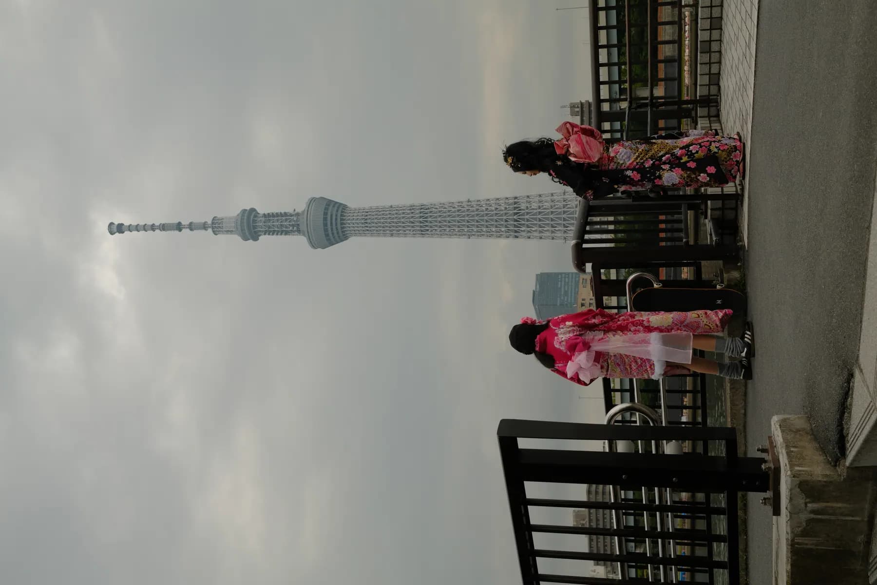 Two women in traditional kimonos stand on a bridge overlooking the Tokyo Skytree on a cloudy day