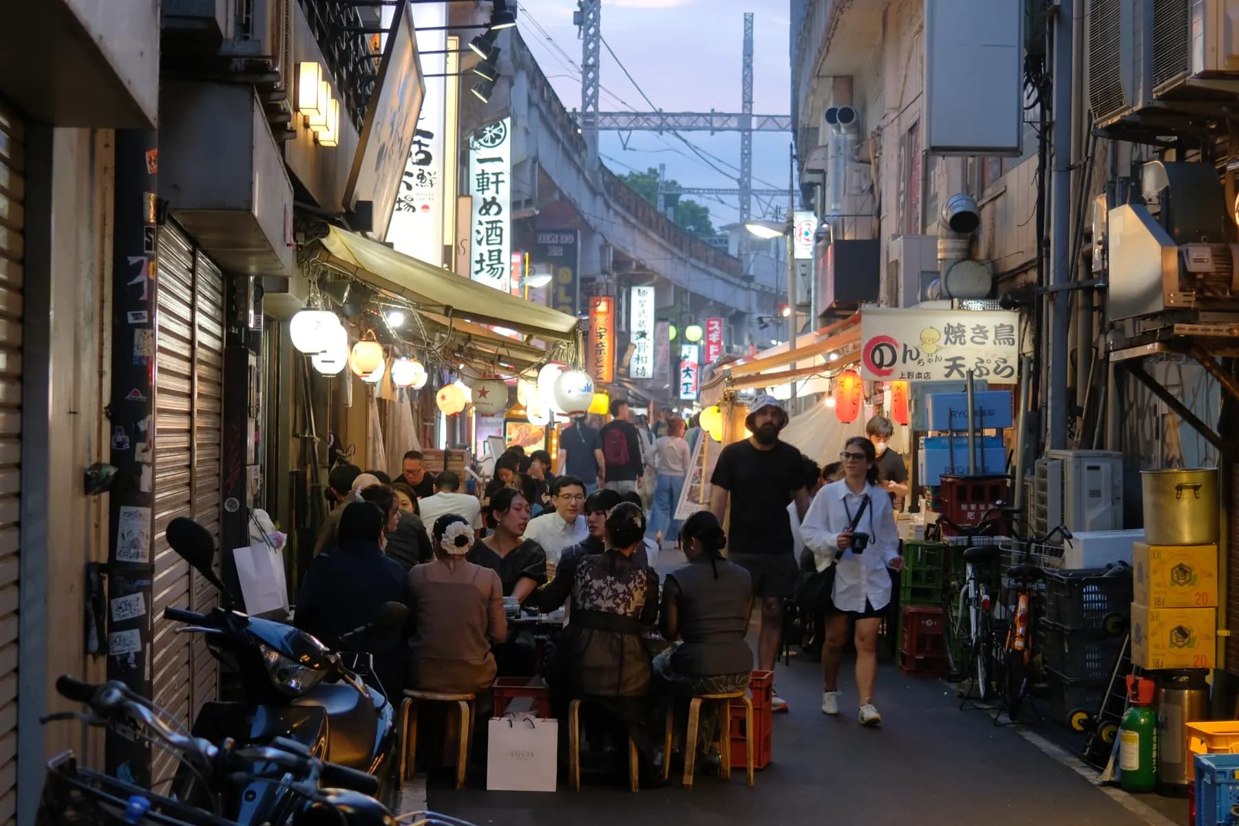 A bustling street scene with people walking and sitting at tables under lanterns, surrounded by various signs and urban elements