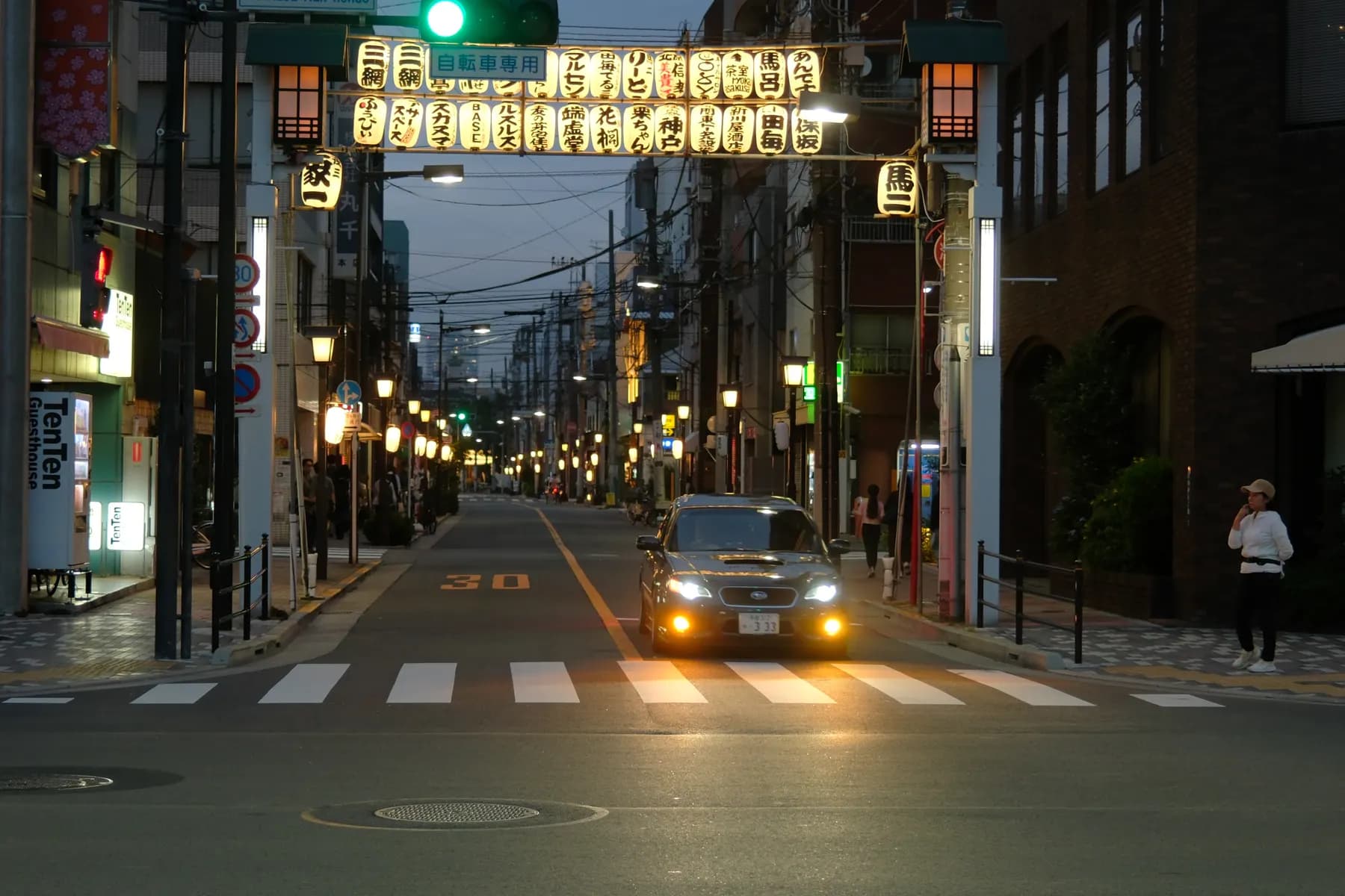 A quiet street scene at night with a car and a pedestrian, illuminated by street lamps and building lights