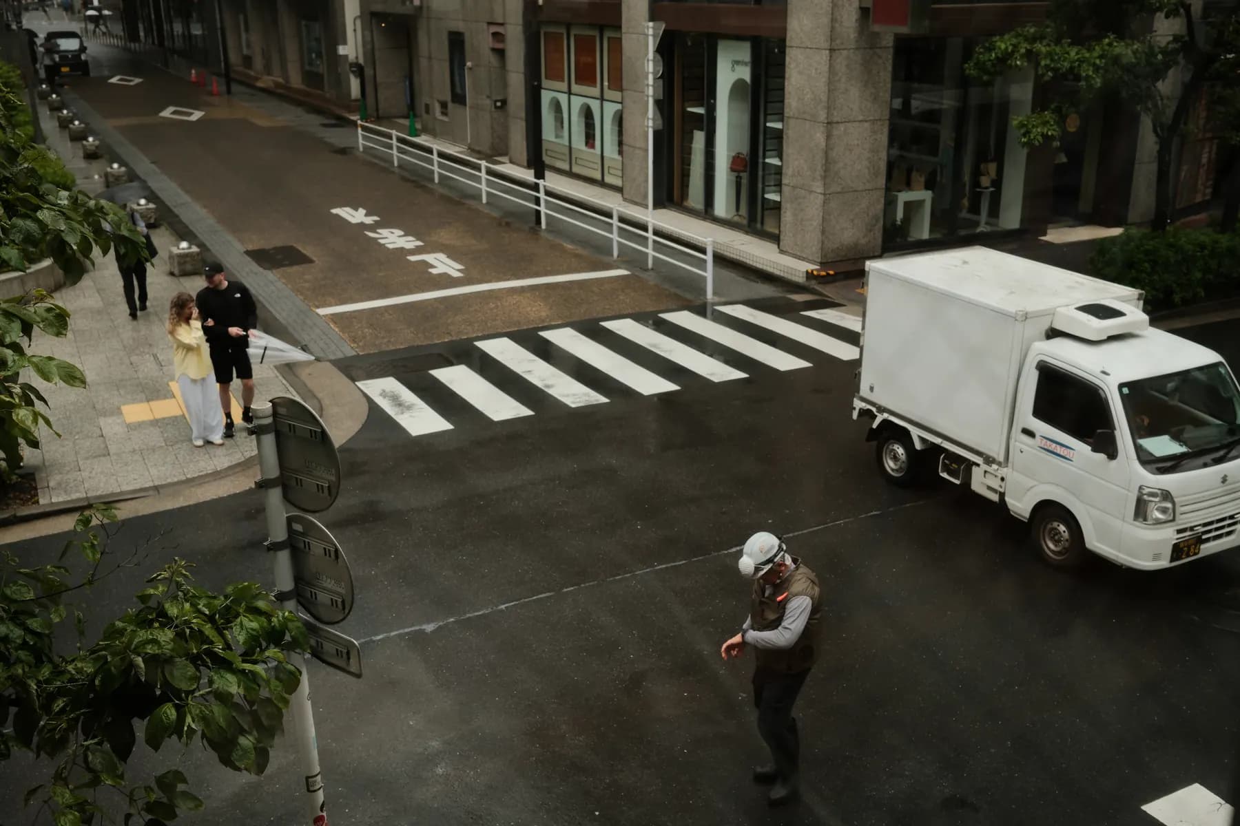 A rainy day street scene with pedestrians, a truck, and a person in a hard hat walking across a crosswalk