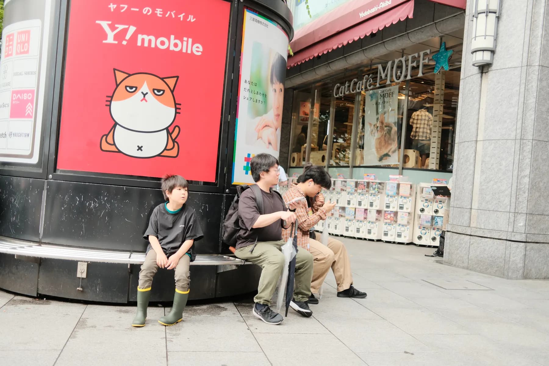 A casual street scene with people sitting on a bench near a Y!mobile advertisement and a cat cafe