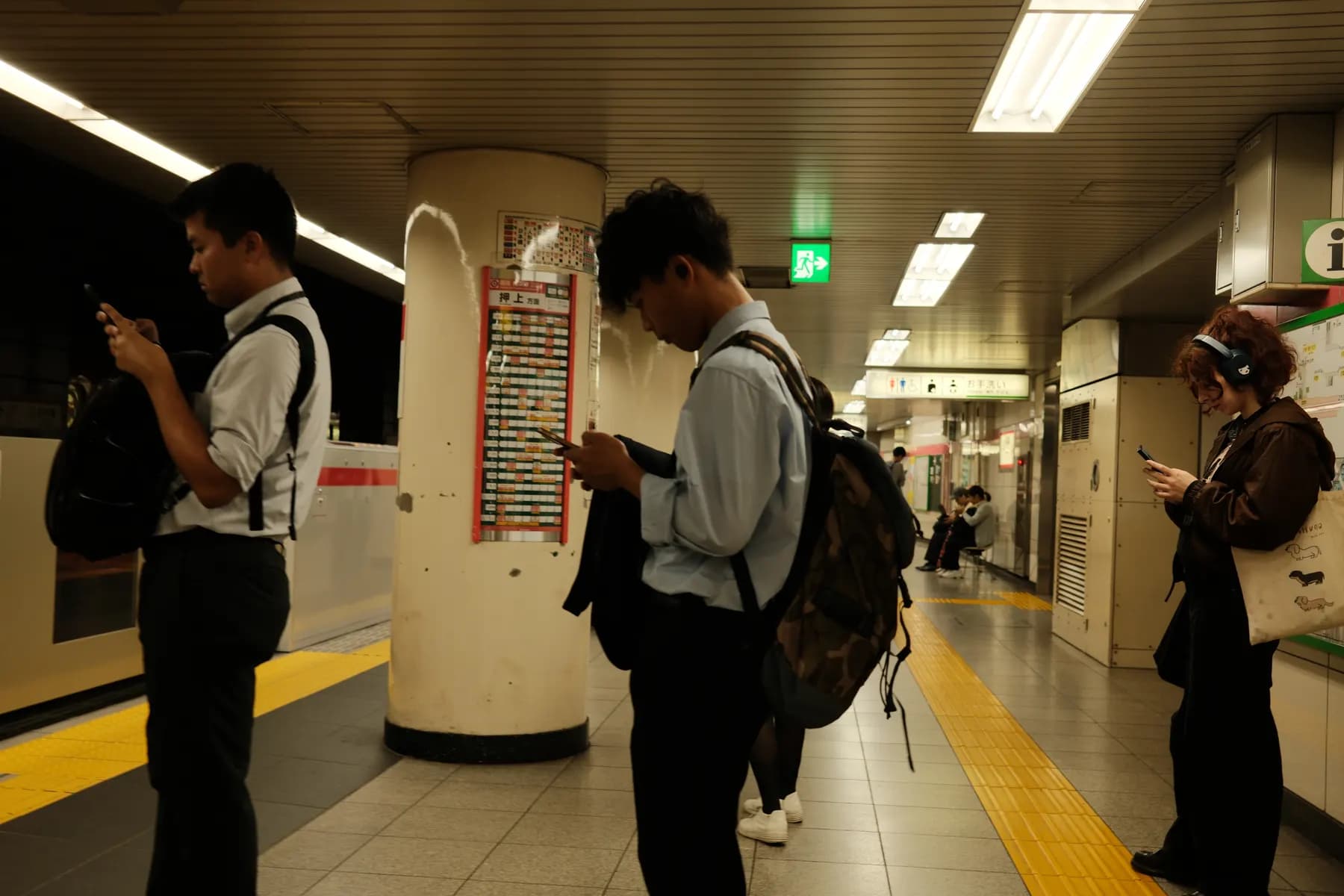 Three individuals are engrossed in their phones in a dimly lit indoor setting, possibly a train station platform, with a poster and a pillar visible in the background