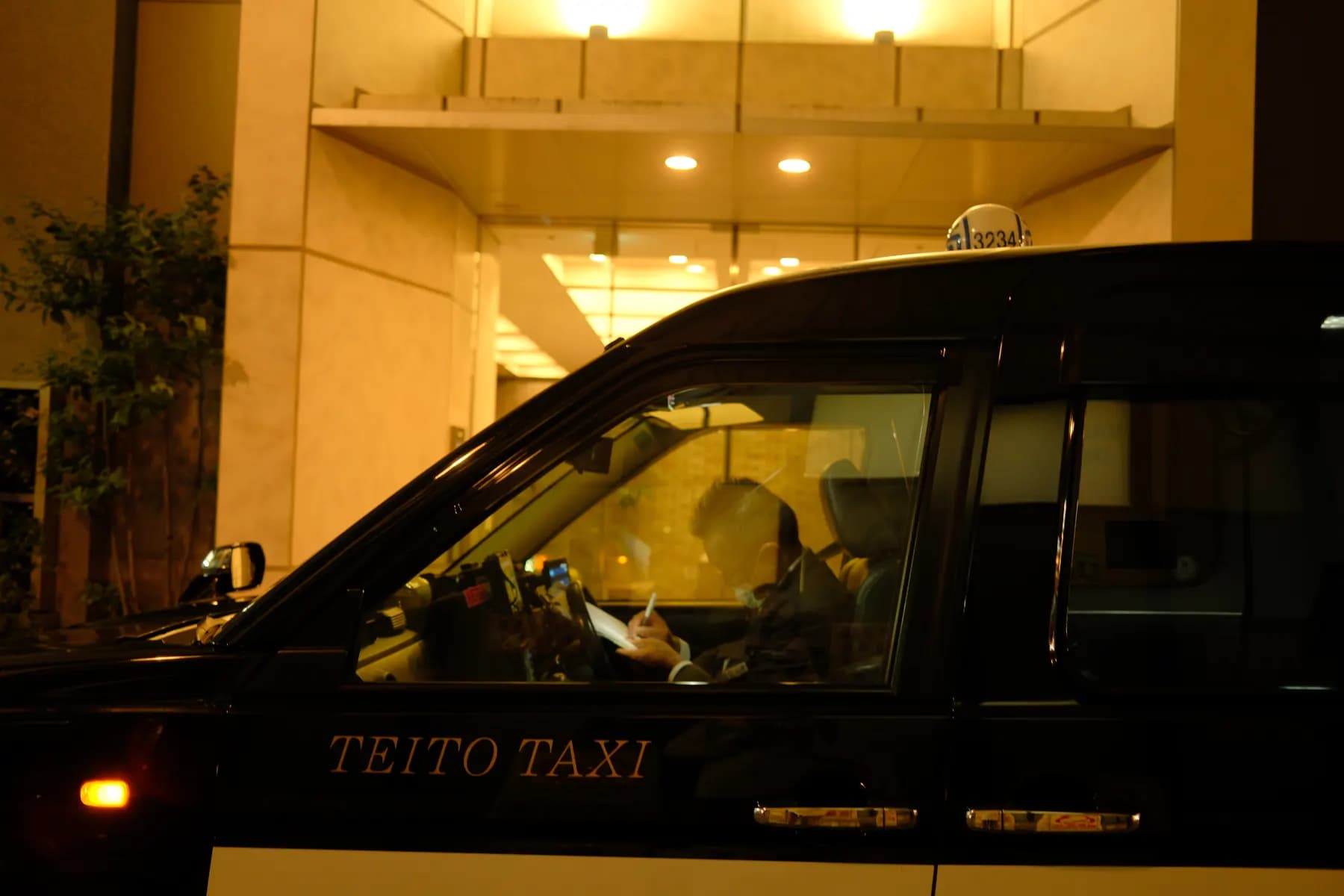 A taxi driver is writing in a notebook while sitting inside a parked taxi at night