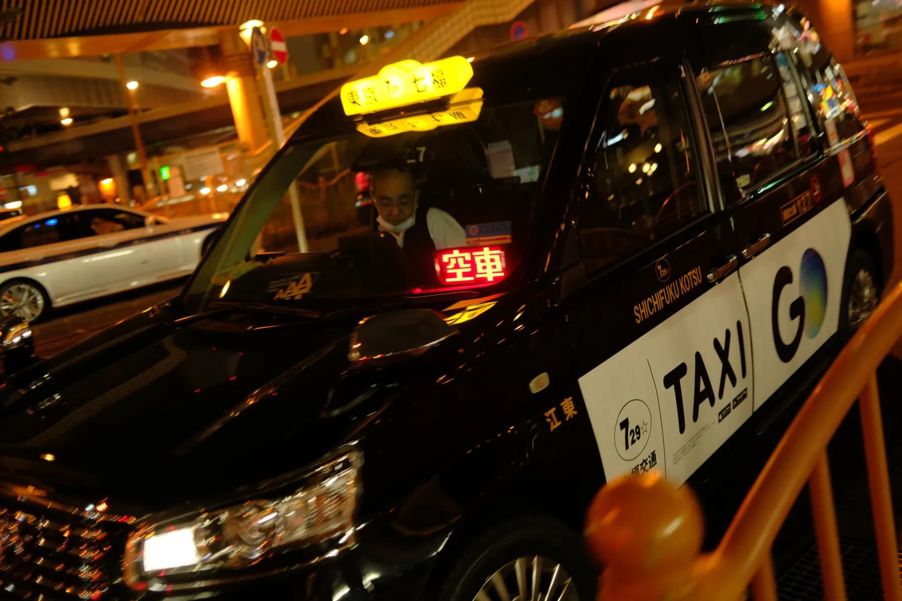 A nighttime urban scene featuring a taxi with a driver inside, illuminated by streetlights and the glow of the taxi's sign
