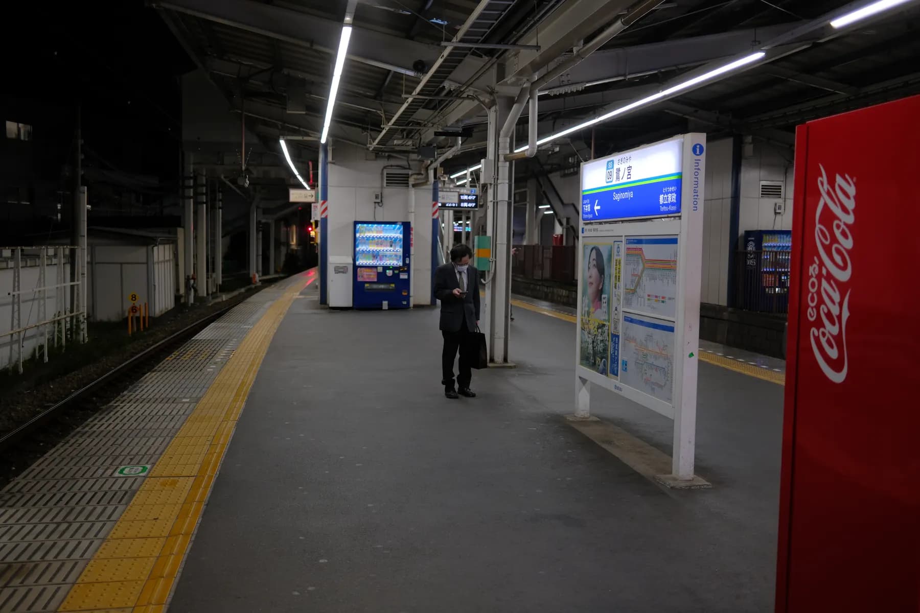 A man in a suit stands on a train platform, looking at his phone, surrounded by urban infrastructure and signage