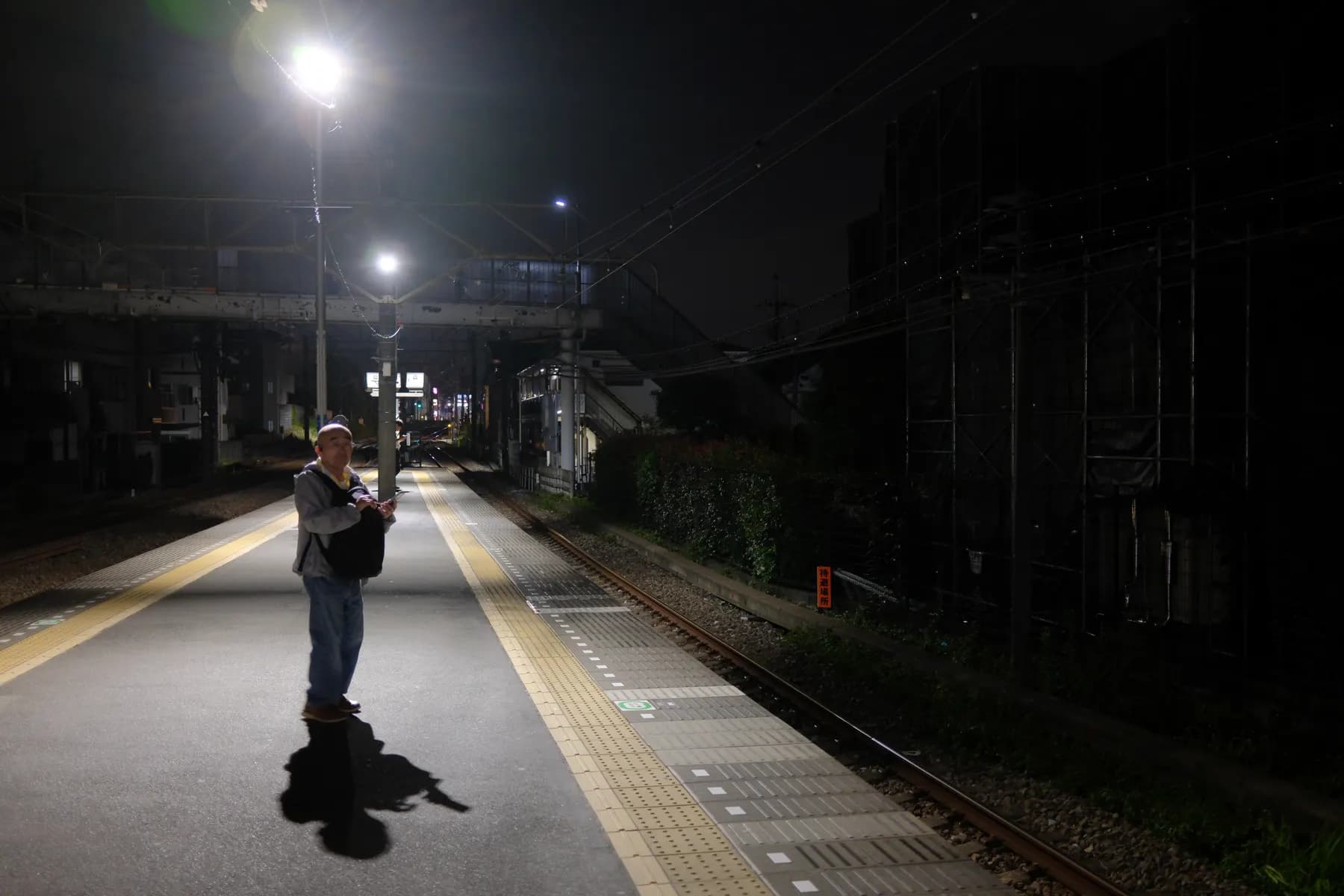 A solitary figure stands on a dimly lit train platform at night, surrounded by urban elements