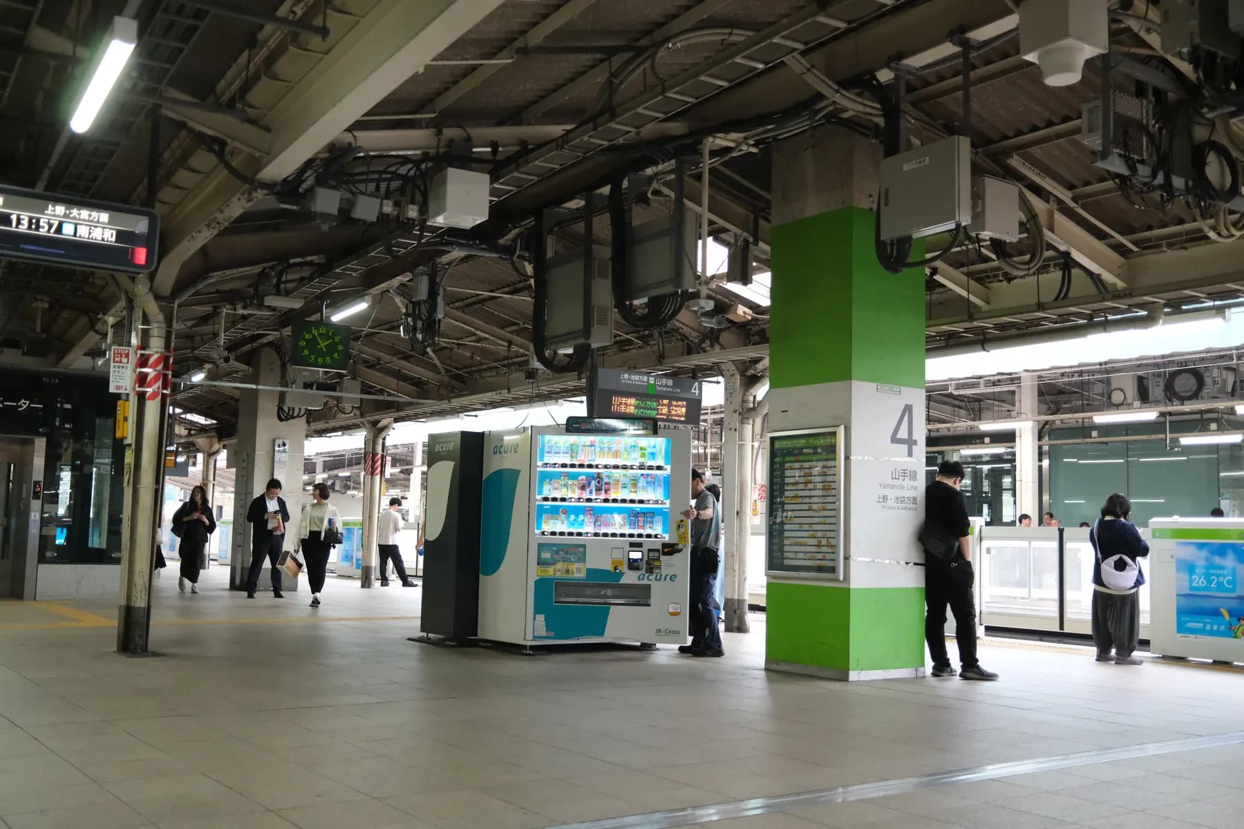 A busy train station platform with people walking and waiting, a vending machine, and various signs and clocks