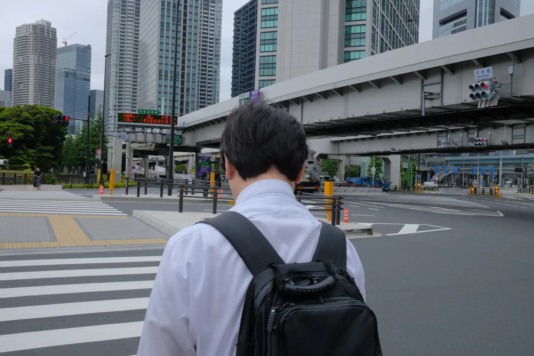 A man in a white shirt and backpack stands at a pedestrian crossing in an urban setting with tall buildings and a bridge in the background
