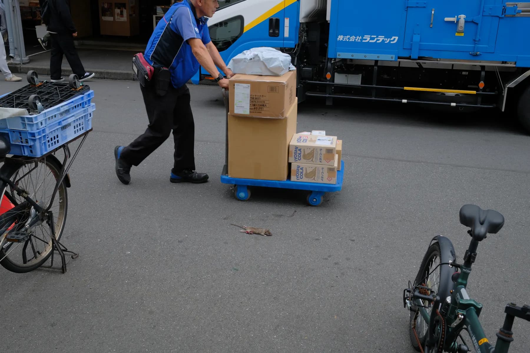 A man in a blue uniform pushes a cart loaded with boxes, while a dead rat lies on the street in front of him