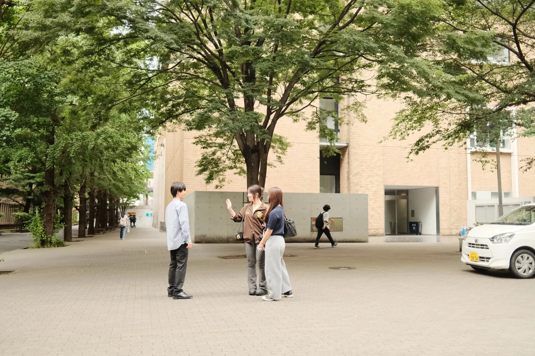 A group of people interacting on a sidewalk in front of a building with trees and a car in the background
