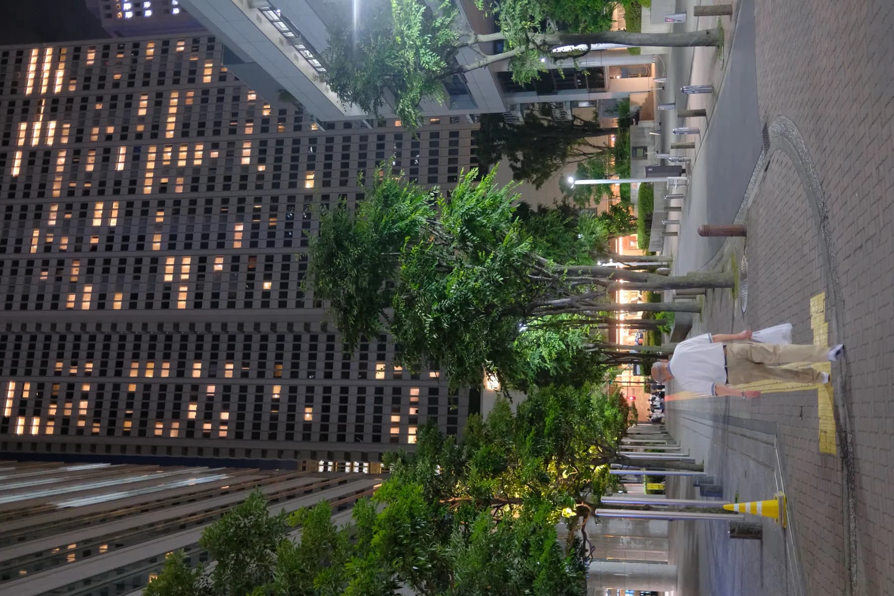 A man walks down a quiet urban street at night, surrounded by tall buildings and illuminated trees