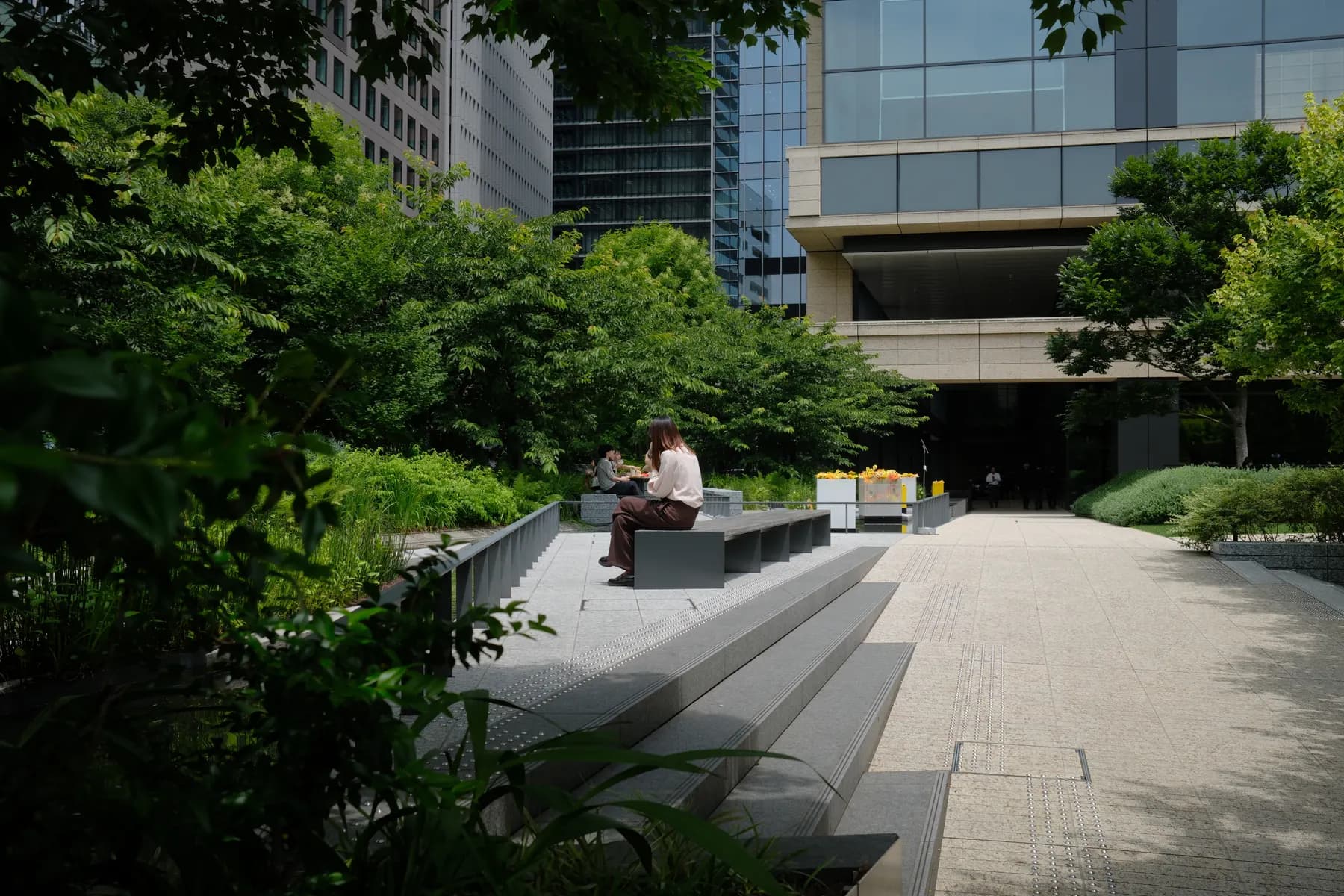 A woman sits on a bench in a serene urban green space surrounded by trees and modern buildings