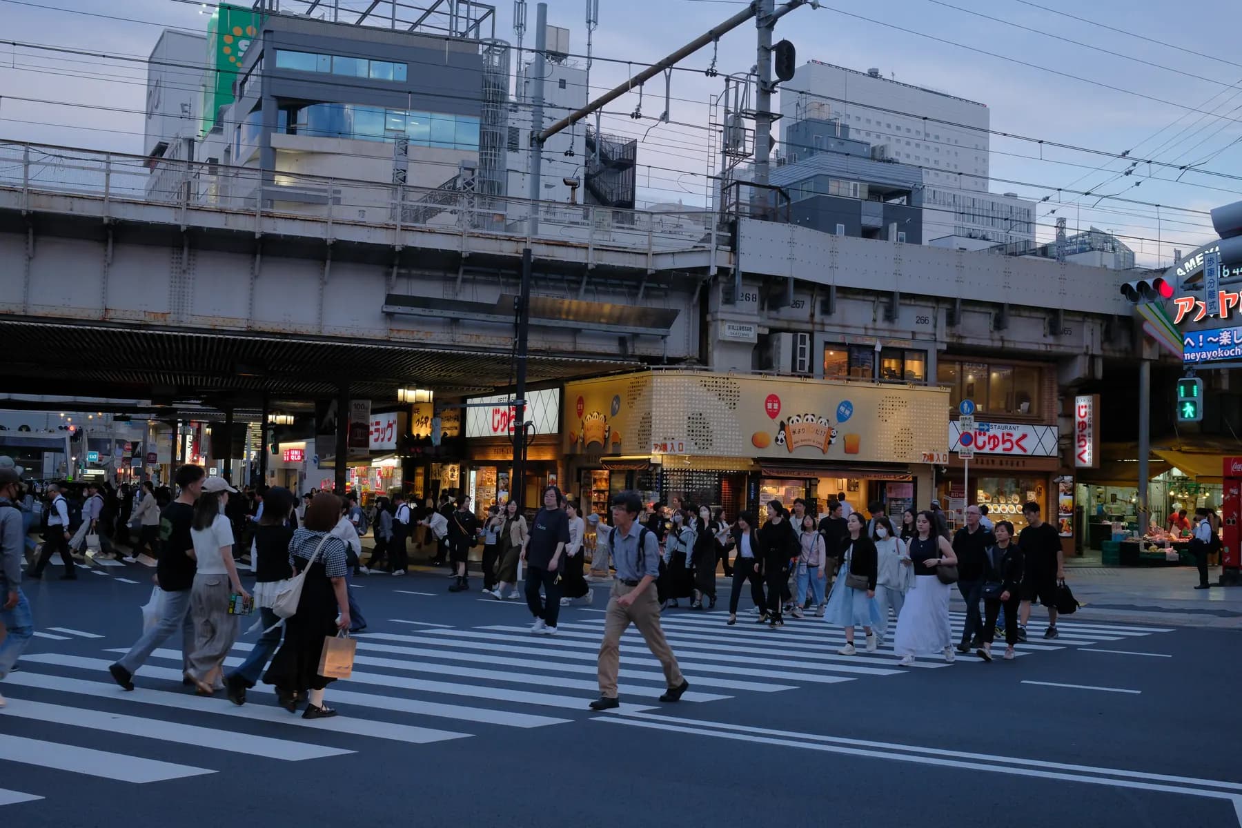 A bustling urban street scene at sunset with pedestrians crossing a crosswalk and various storefronts and signage in the background