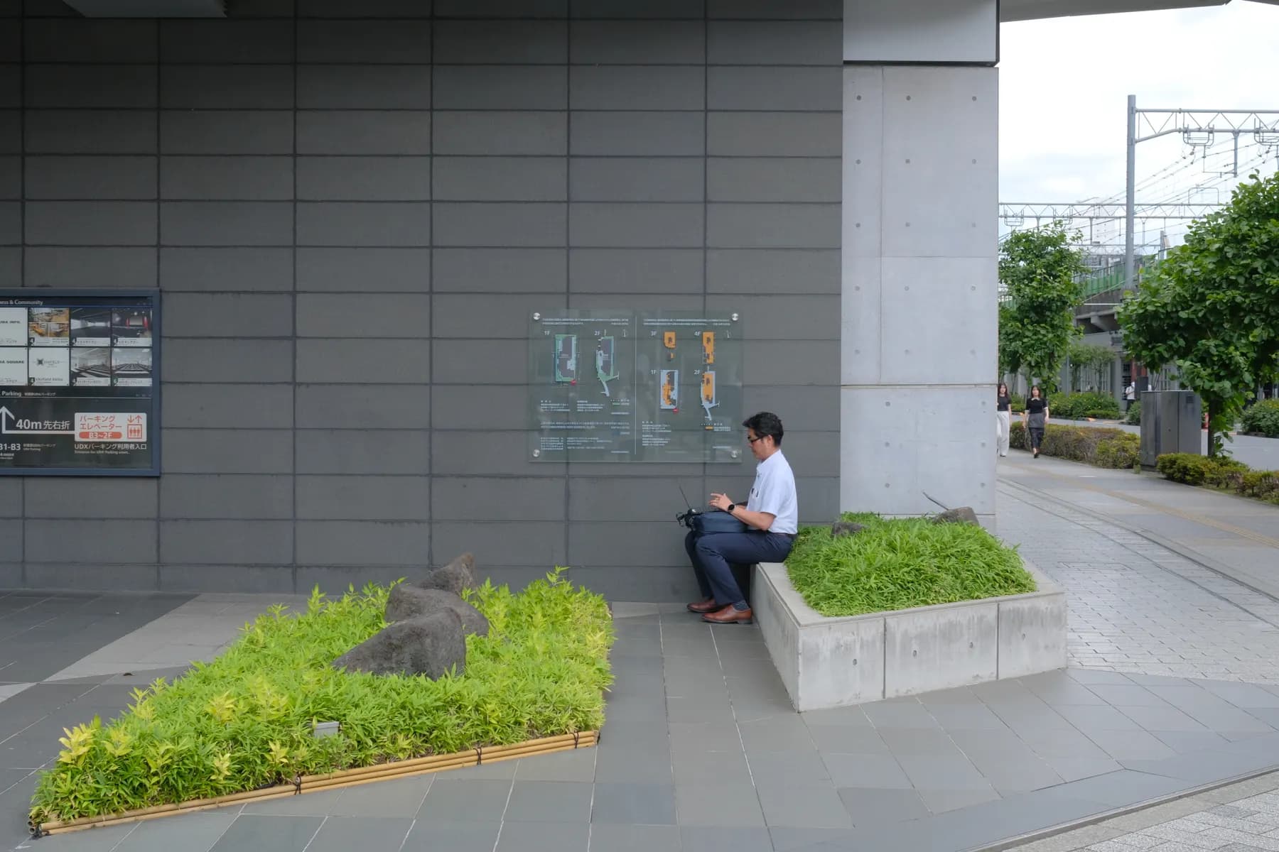 A man is sitting on a bench in an urban setting, using a laptop, surrounded by greenery and informational signs