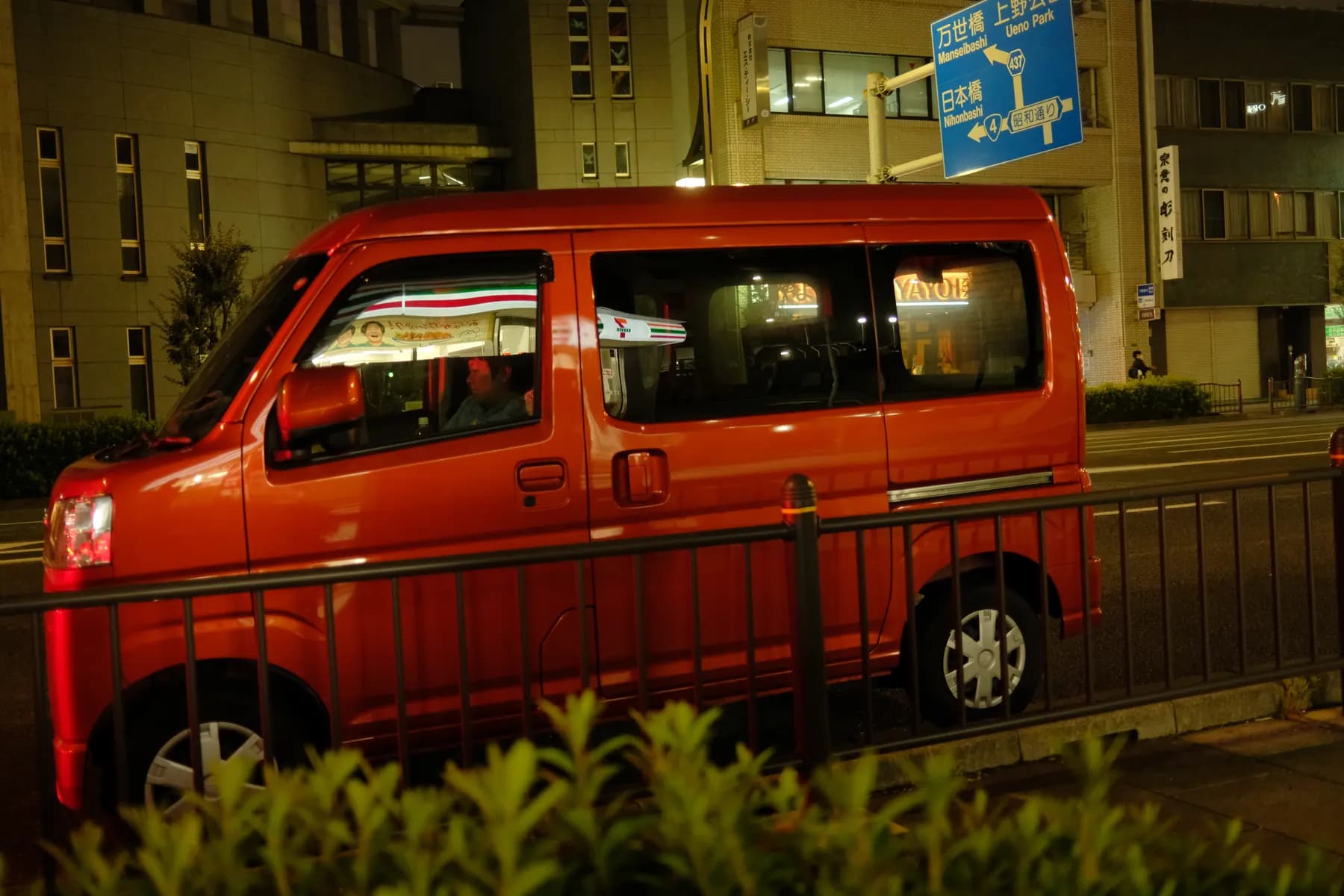 A red van is parked on a street at night, with a street sign and urban buildings in the background
