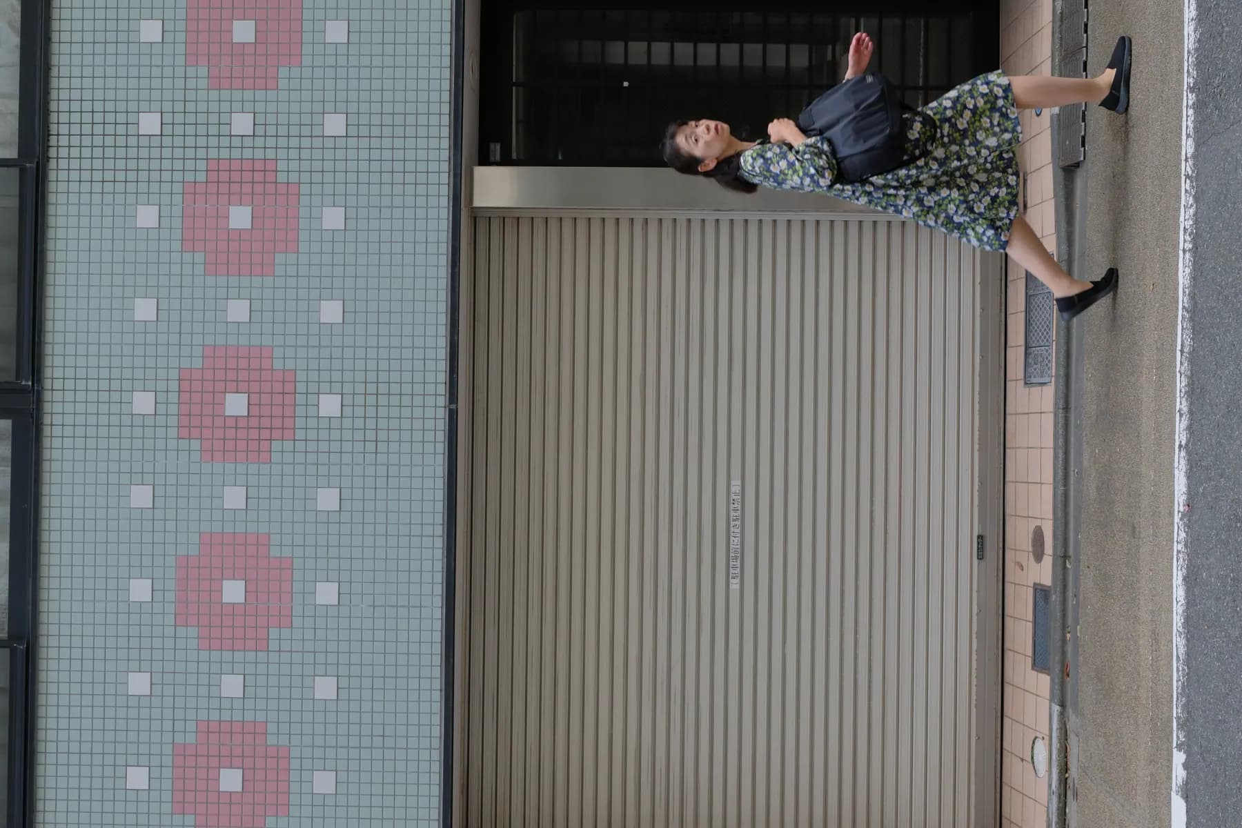 A woman in a floral dress walks past a shuttered storefront with a tiled facade in an urban setting